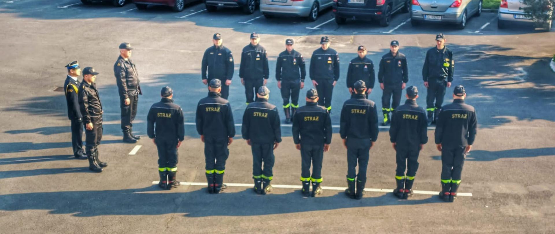 Obraz przedstawia strażaków jaworskiej komendy podczas zmiany służby. Strażacy stoją naprzeciw siebie w dwóch szeregach. W zmianie służby uczestniczy Komendant Powiatowy oraz dowództwo JRG Jawor