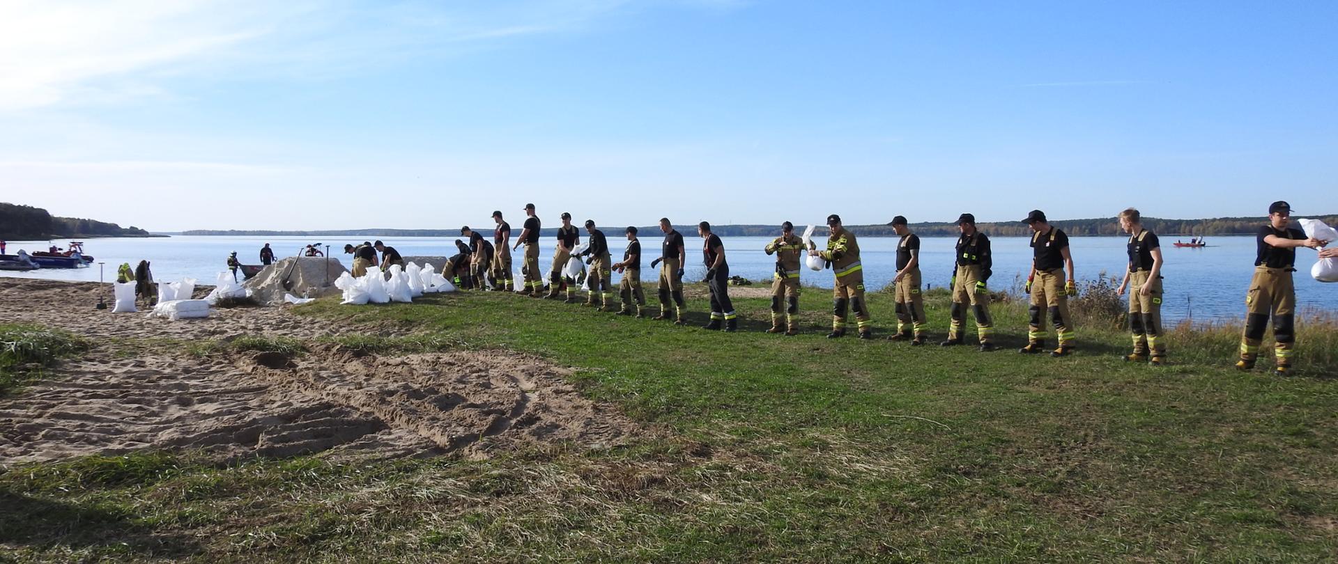 Zdjęcie przedstawia strażaków stojących w rzędzie i podających z rąk do rąk worki z piaskiem. W tle widać zbiornik wodny.