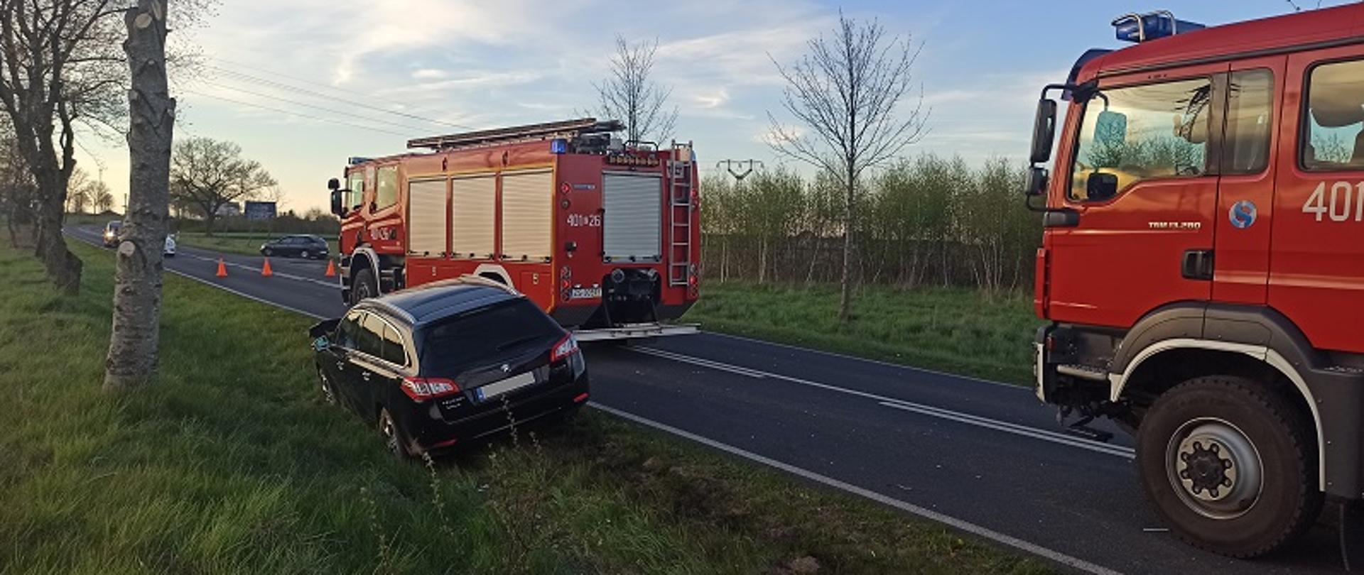 Zdjęcie przedstawia stojący na poboczu widoczny od tyłu czarny samochód marki Peugeot. Po prawej stronie widoczne od strony kierowcy stojące na drodze dwa samochody pożarnicze