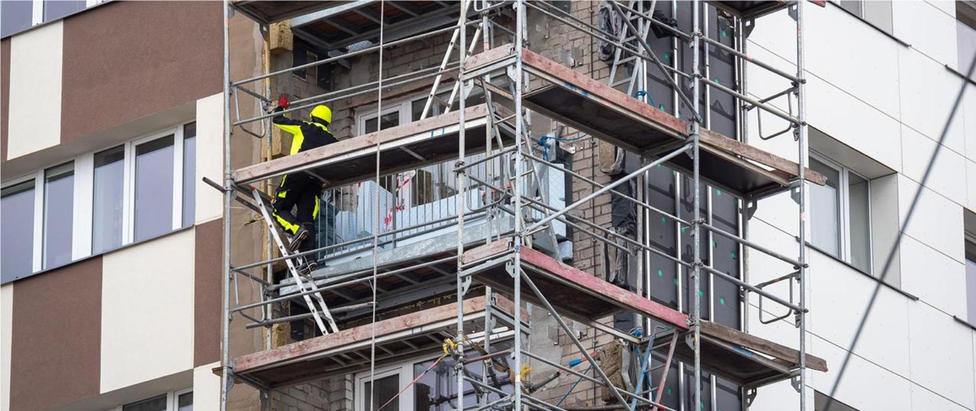 narożnik bloku w trakcie termomodernizacji - z lewej gotowa elewacja, na środku balkony i ściana z cegły, rusztowanie, na nim robotnik budowlany w kasku i odzieży ochronnej, z prawej widoczne warstwy nowej elewacji