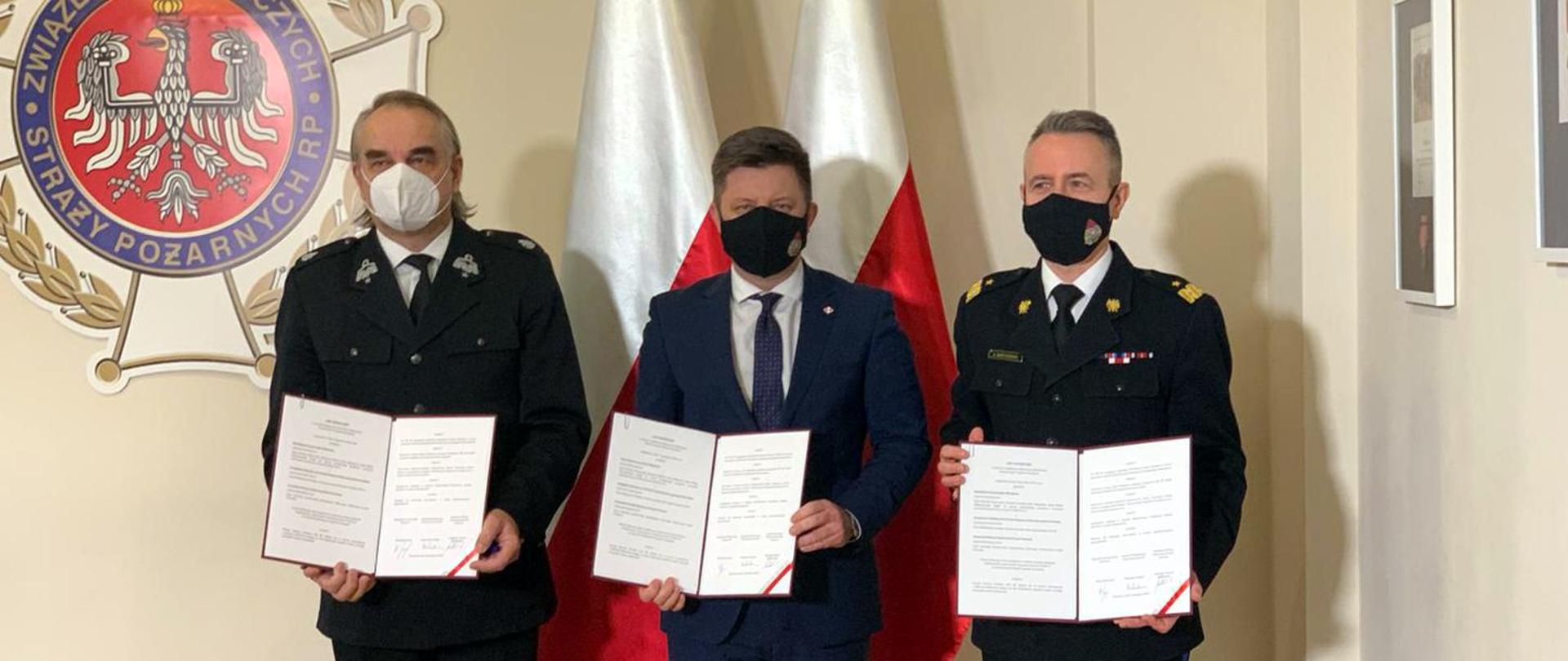 Siedziba Zarządu Głównego Związku Ochotniczych Straży Pożarnych Rzeczypospolitej Polskiej. Na tle dwóch biało-czerwonych flag i wiszącego na ścianie herbu Związku Ochotniczych Straży Pożarnych stoją: szefa Kancelarii Prezesa Rady Ministrów - Michał Dworczyk, komendant główny PSP - nadbryg. Andrzej Bartkowiak i prezes ZG ZOSP RP - dh Waldemar Pawlak. W rękach trzymają podpisany list intencyjny w sprawie współpracy dotyczącej realizowania Narodowego Programu Szczepień.