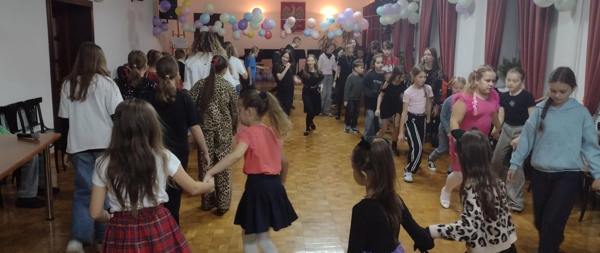 Na zdjęciu widać dzieci, którzy biorą udział w zabawie andrzejkowej. Dzieci dobrane są w pary i tańczą. Sala koncertowa jest udekorowana kolorowymi balonami.