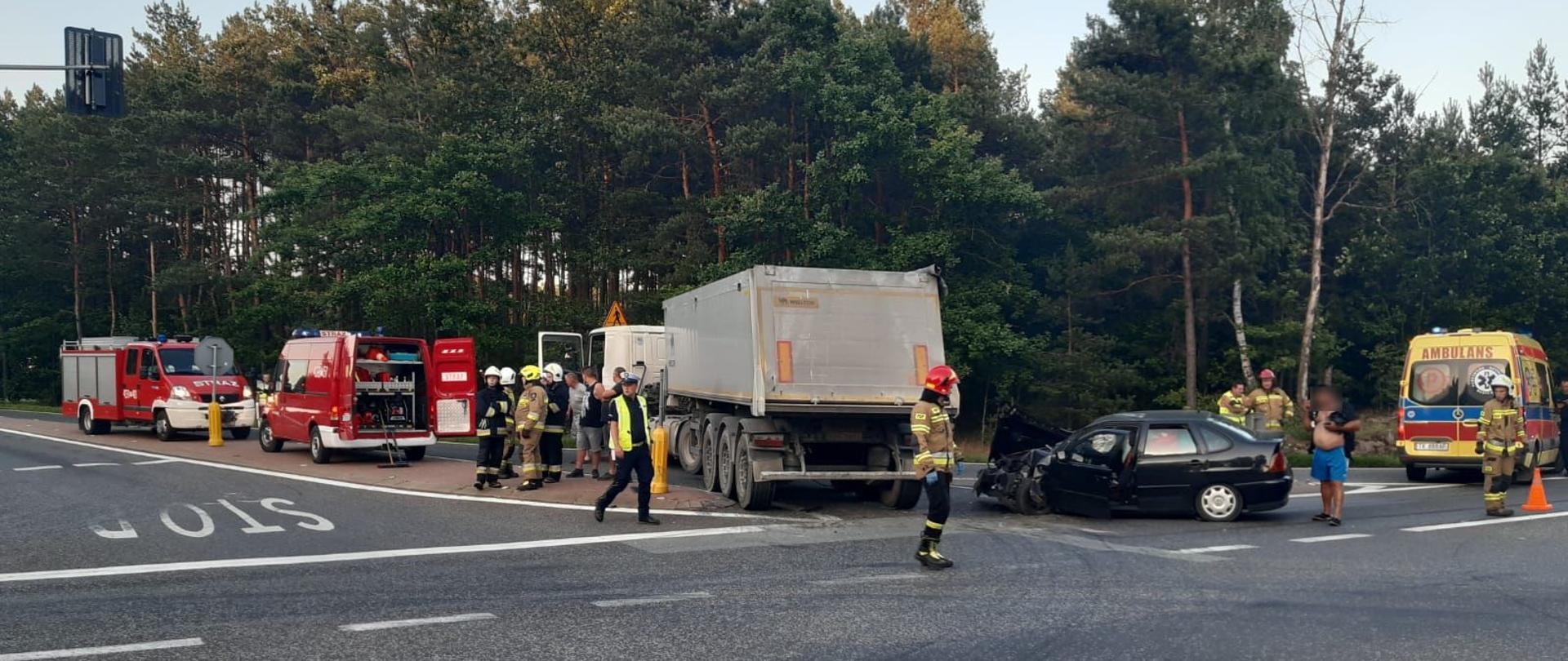 Służby prowadzą czynności przy wypadku, ciężarówka skręciła w lewo, w jej bok uderzyła osobówka VW Polo która ma rozbity przód. Po lewej stronie ciężarówki dwa samochody strażackie. Po prawej od strony samochodu osobowego stoi karetka.