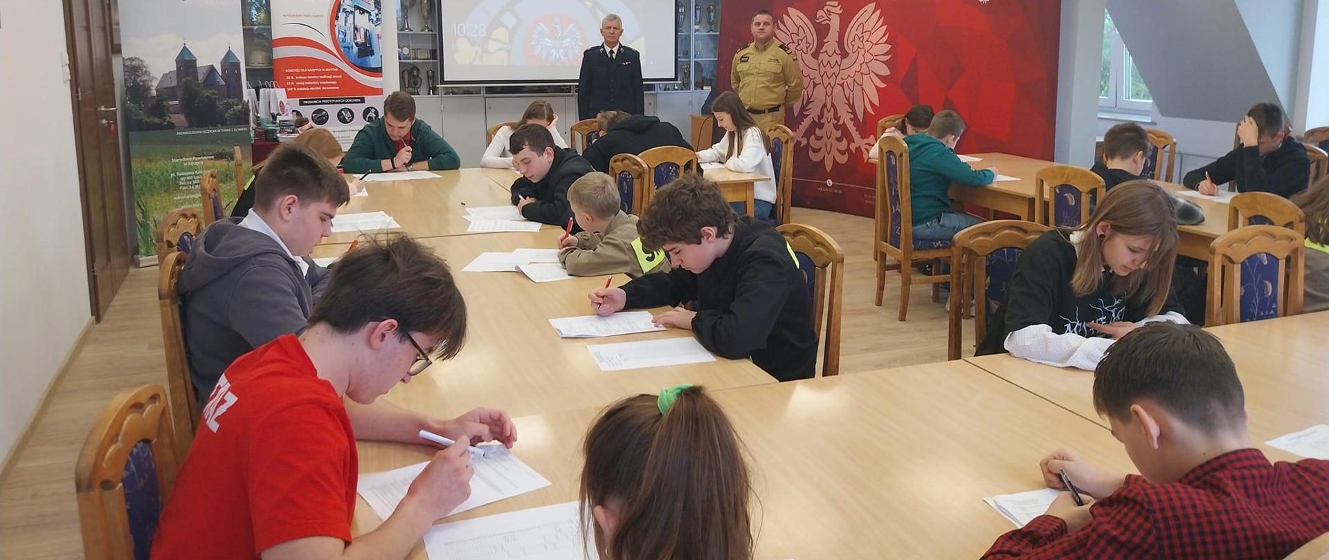 Na zdjęciu widać dwóch strażaków. Jeden jest w żółtym stroju a drugi w ciemnym. Dzieci siedzą przy stole i rozwiązują test W tle widać obraz na ścianie i banery reklamowe. Wydarzenie odbywa się wewnątrz budynku. Jest widno.