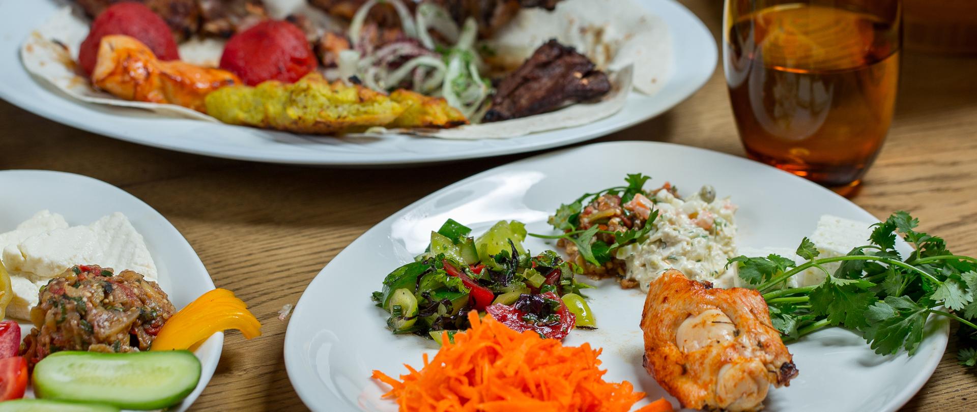 Na pierwszym planie, na dole zdjęcia po prawej stronie, widać w dużym przybliżeniu talerzyk z ułożonymi oddzielnie porcjami pokrojonej w słupki marchewki, zielonych sałat z sosem, upieczonego kurczaka oraz kilku gałązek świeżej zielonej pietruszki. Nad talerzykiem po prawej stronie, widoczna brązowa przezroczysta szklanka z wodą. Po lewej stronie na dole zdjęcia, widać część białego talerzyka z ułożonymi dwoma plastrami ogórka, kawałkiem żółtej papryki, porcją sałatki i dwoma plastrami twarogu. Po lewej stronie na górze zdjęcia, widać duży biały głęboki talerz z płatem tortilli i ułożonymi na nim kilkoma porcjami pieczonego mięsa, dwoma czerwonymi małymi pomidorkami i ogórkiem pokrojonym w sprężynkę. Nad talerzem widać nóź w dłoni oraz widelec sięgające po mięso z talerza. W górnej części zdjęcia w tle widać dość niewyraźnie części białych talerzyków z masłem, warzywami, dwa małe białe pojemniczki oraz część koszyka z pieczywem. Wszystko ustawione jest na drewnianym stole.