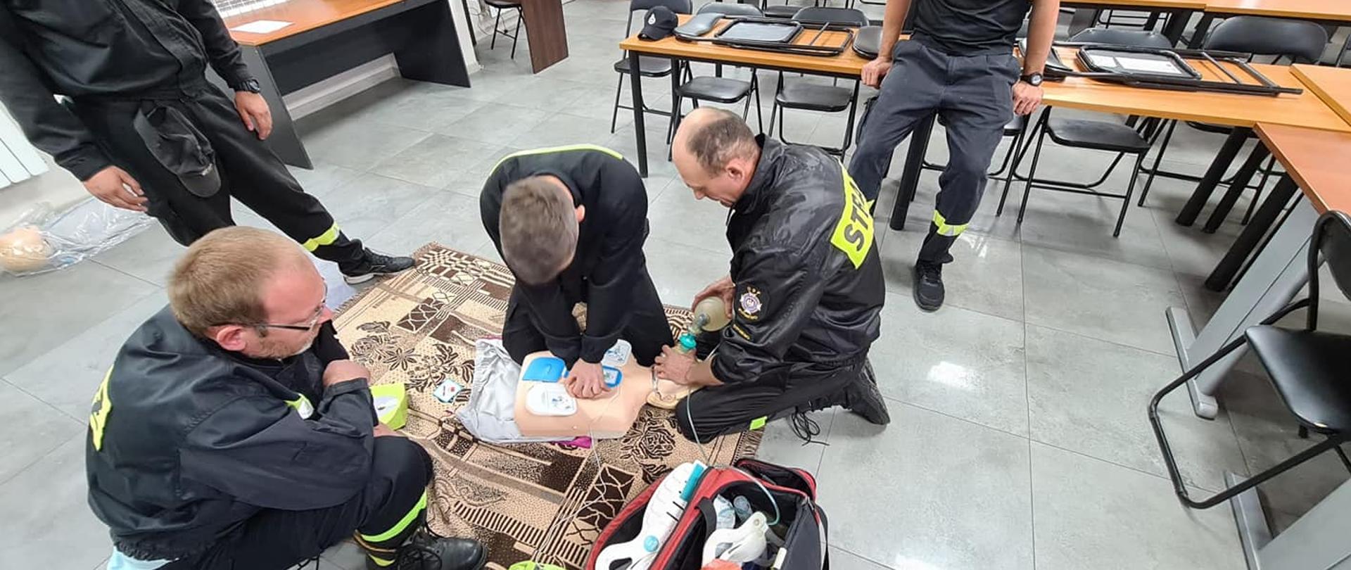 Trzech strażaków udziela kwalifikowanej pierwszej pomocy manekinowi leżącemu na podłodze na kocu. Pierwszy z nich uciska klatkę piersiową, na której przyklejono klatkę piersiową. Drugi podaje tlen przy pomocy worka ambu. Trzeci klęczy przy nich. 