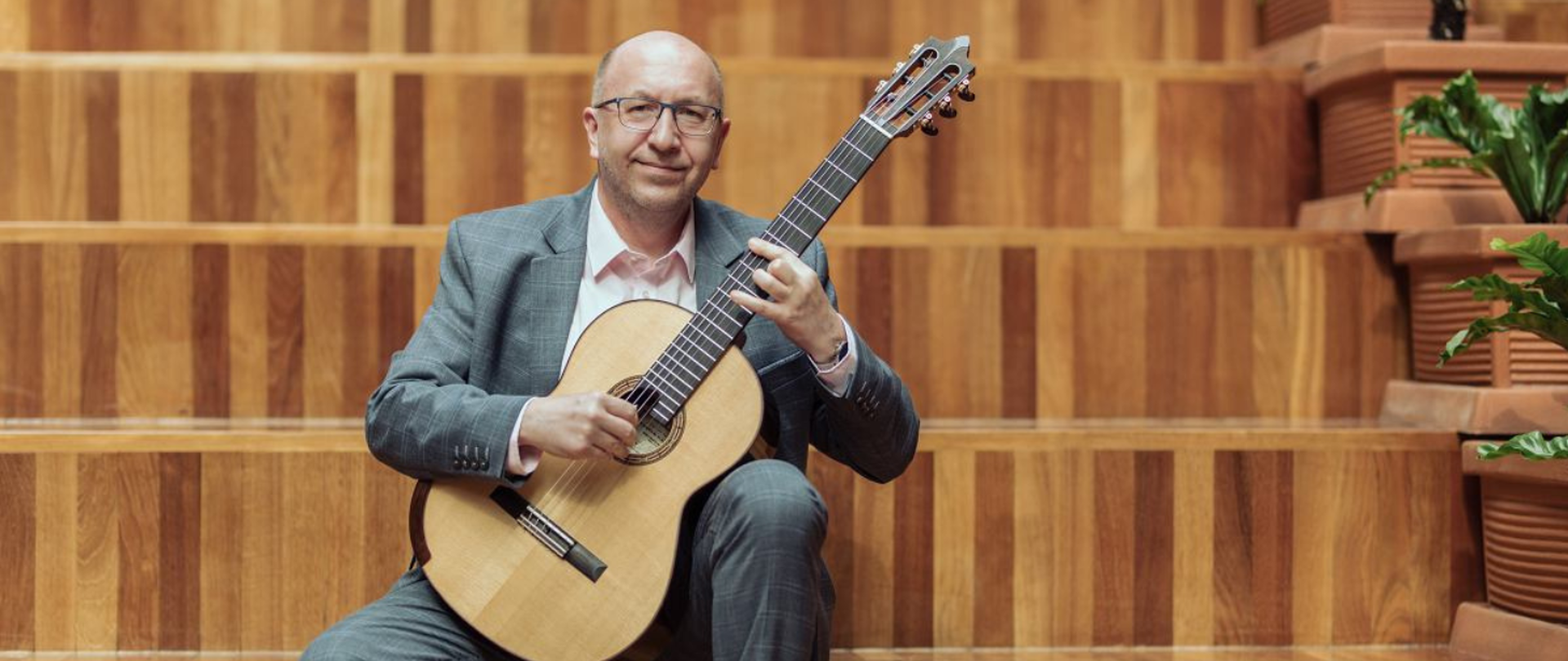 Na zdjęciu widać mężczyznę w szarym garniturze siedzącego na drewnianych schodach i grającego na gitarze klasycznej. W tle drewniana ściana i rośliny.