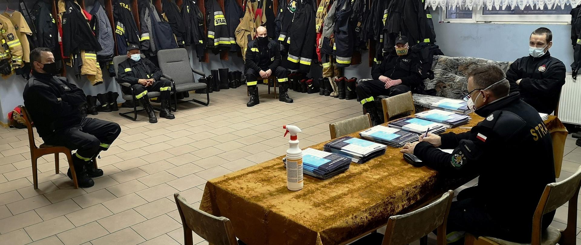 Widok pomieszczenia remizy OSP w Cybince. Na krzesłach w oddaleniu od siebie siedzą umundurowani strażacy OSP. Za stołem, na którym leżą ulotki, siedzi Komendant Gminny ZOSP RP w Cybince.