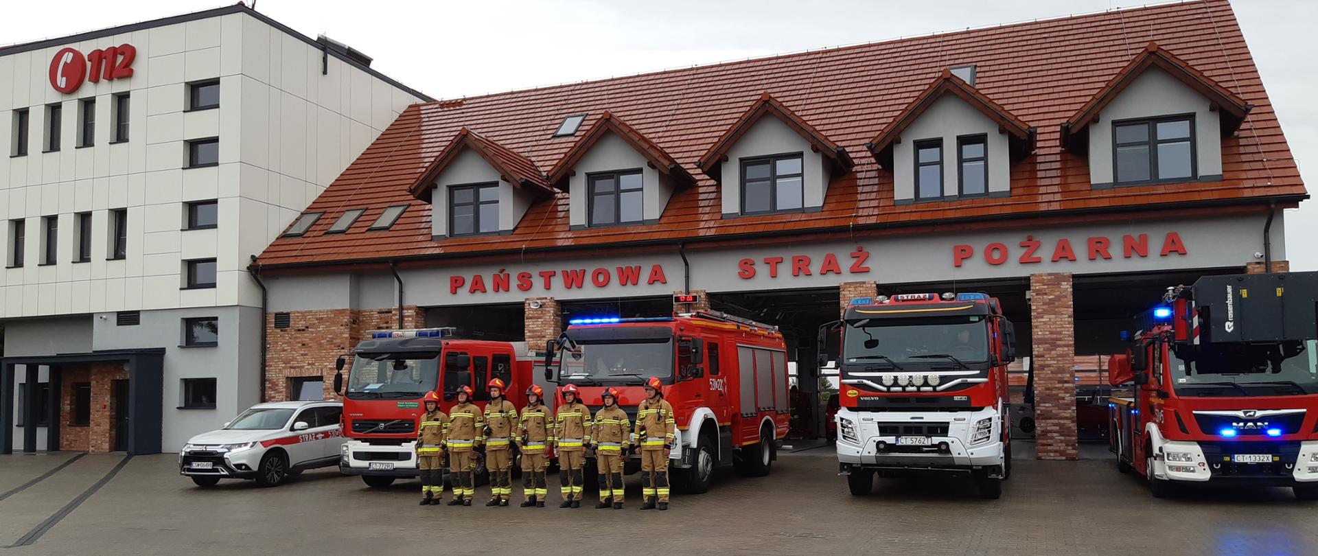Przed budynkiem Komendy Powiatowej Państwowej Straży Pożarnej w Świeciu stoją samochody pożarnicze z włączonymi sygnałami dźwiękowymi oraz błyskowymi. Przed pojazdami stoją funkcjonariusze ubrani w ubrania specjalne i hełmy. 
