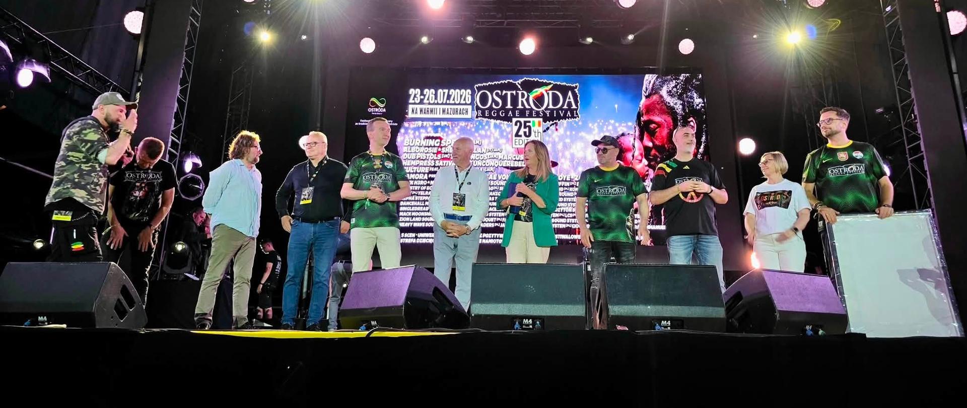 Grupa oficjeli stoi na dużej scenie Ostróda Reggae Festival. W tle widoczny baner festiwalu oraz program wydarzenia