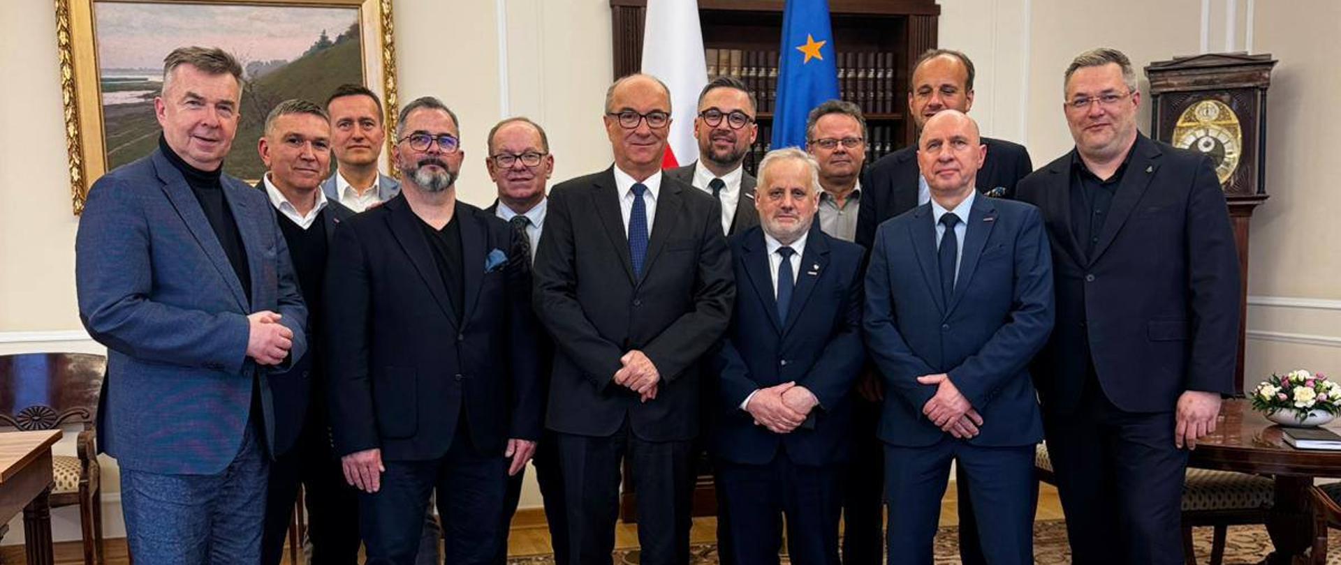 mężczyźni w garniturach stoją na tle flagi Polski i UE, pozują do zdjęcia