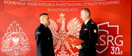 Na zdjęciu widać dwóch mężczyzn w wyjściowych mundurach straży pożarnej, stojących naprzeciwko siebie. Jeden z nich - Śląski Komendant Wojewódzki PSP - st. bryg. Damian Legierski trzyma w rękach czerwony dokument – decyzję o powierzeniu obowiązków, który wręcza kpt. Krzysztofowi Sadowskiemu, pełniącemu obowiązki zastępcy komendanta powiatowego PSP w Będzinie. Tło zdjęcia jest intensywnie czerwone, z dużym herbem Polski (biały orzeł) pośrodku. Na górze tła widnieje biały napis: „PAŃSTWOWA STRAŻ POŻARNA KOMENDA WOJEWÓDZKA PAŃSTWOWEJ STRAŻY POŻARNEJ W KATOWICACH”. Po lewej stronie w tle znajduje się stylizowane logo straży pożarnej z orłem w hełmie strażackim. Po prawej stronie widnieje biały napis „KSRG 30 LAT” – oznaczający 30-lecie Krajowego Systemu Ratowniczo-Gaśniczego.