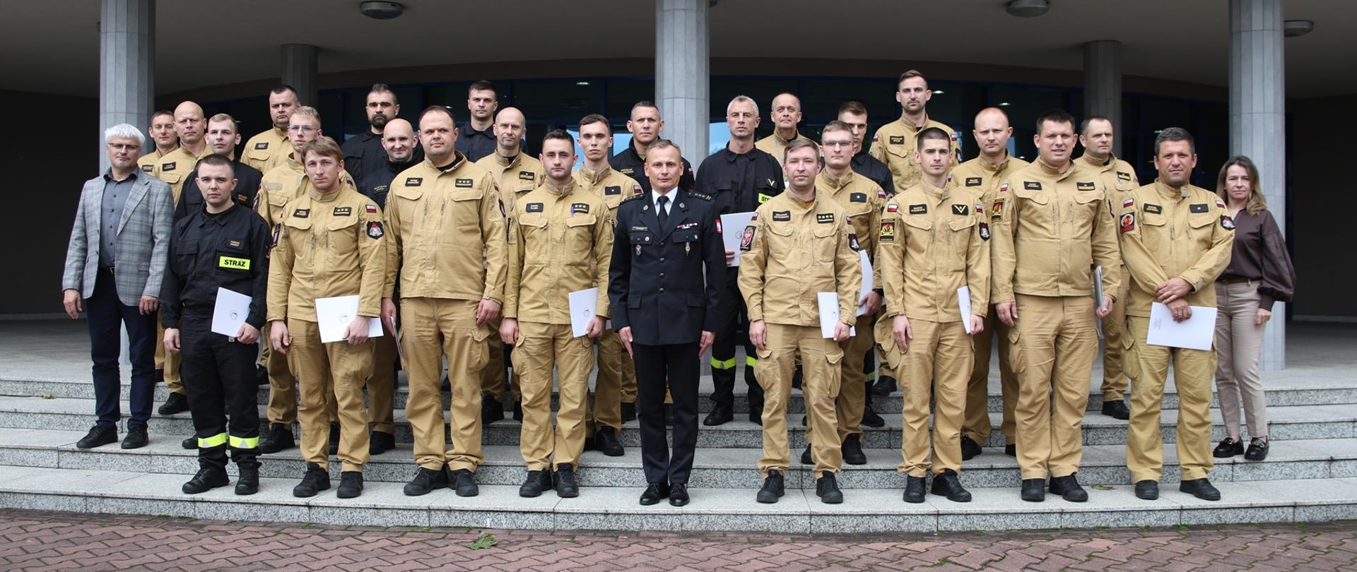 Pamiątkowe zdjęcie grupowe funkcjonariuszy PSP będących uczestnikami szkolenia z zakresu awaryjnego rozładunku cystern (24 osoby) w towarzystwie Zastępcy Komendanta Centralnej Szkoły PSP, kobiety, dwóch funkcjonariuszy CS PSP oraz wykładowcy