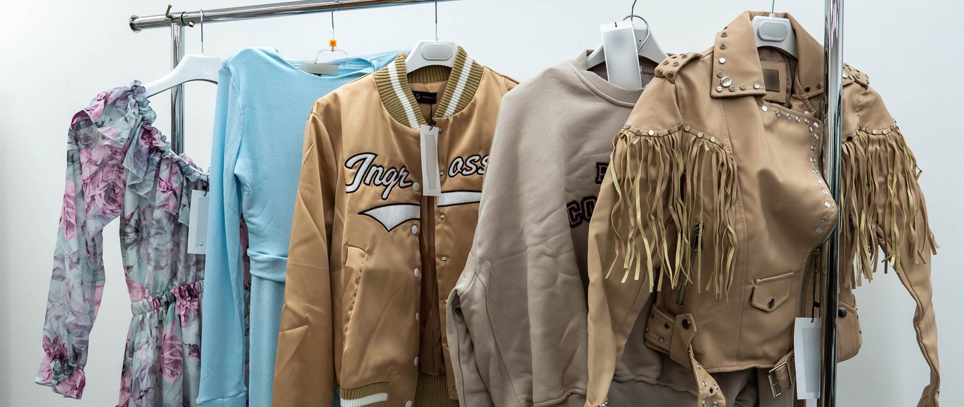 Na wieszaku na tle białej ściany wiszą dwie kurtki, bluza i dwie sukienki.