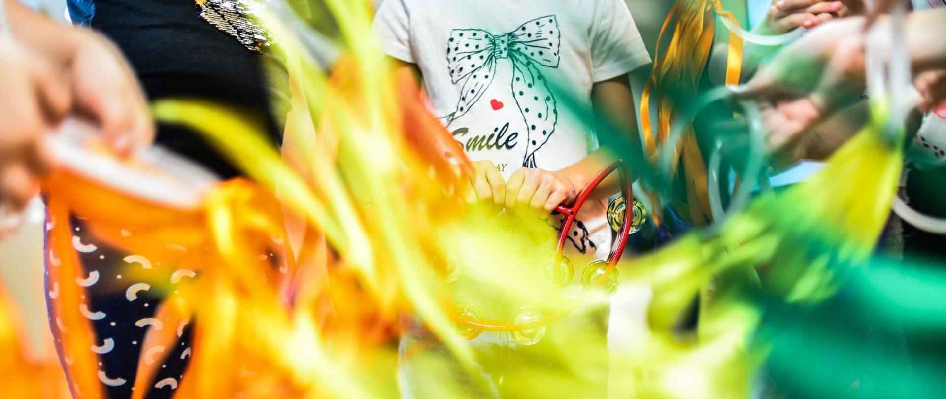 kolorowe zdjęcie przedstawia dzieci bawiące się kolorowymi szarfami, w centrum fotografii dziewczynka w białej koszulce z napisem “smile”