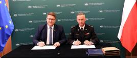 Na zdjęciu widzimy dwóch mężczyzn siedzących przy czarnym stole w Sali konferencyjnej , Wojewódzkiego Funduszu Ochrony Środowiska i Gospodarki Wodnej w Katowicach podpisujących umowy. Po lewej stronie siedzi Prezes Wojewódzkiego Funduszu Ochrony Środowiska i Gospodarki wodnej w Katowicach- Mateusz Pindel, w garniturze z niebieskim krawatem i okularami. Po prawej stronie siedzi Śląski Komendant Wojewódzki PSP st. bryg. Damian Legierski w ciemnym mundurze wyjściowym z odznaczeniami i pagonami oficerskimi. Obaj mają przed sobą otwarte teczki z dokumentami i trzymają długopisy w rękach. Na stole leżą także dodatkowe teczki i kilka długopisów. W tle znajduje się zielona ścianka z białymi napisami, na której wielokrotnie powtórzono nazwę : „Wojewódzki Fundusz Ochrony Środowiska i Gospodarki Wodnej w Katowicach”. Po lewej stronie widoczna jest flaga Unii Europejskiej, a po prawej – flaga Polski.