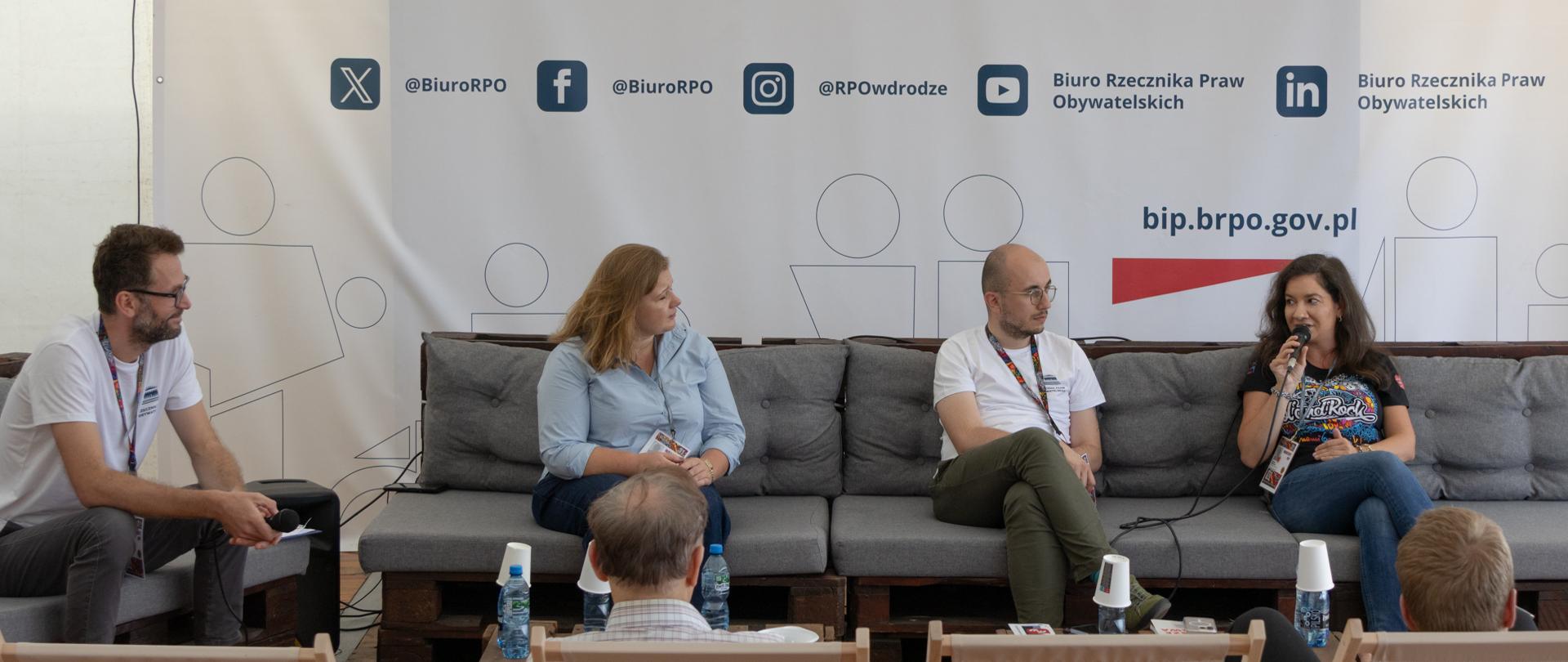 Cztery osoby rozmawiają siedząc na kanapie. Przed kanapami widać leżaki, na których siedzi publiczność.