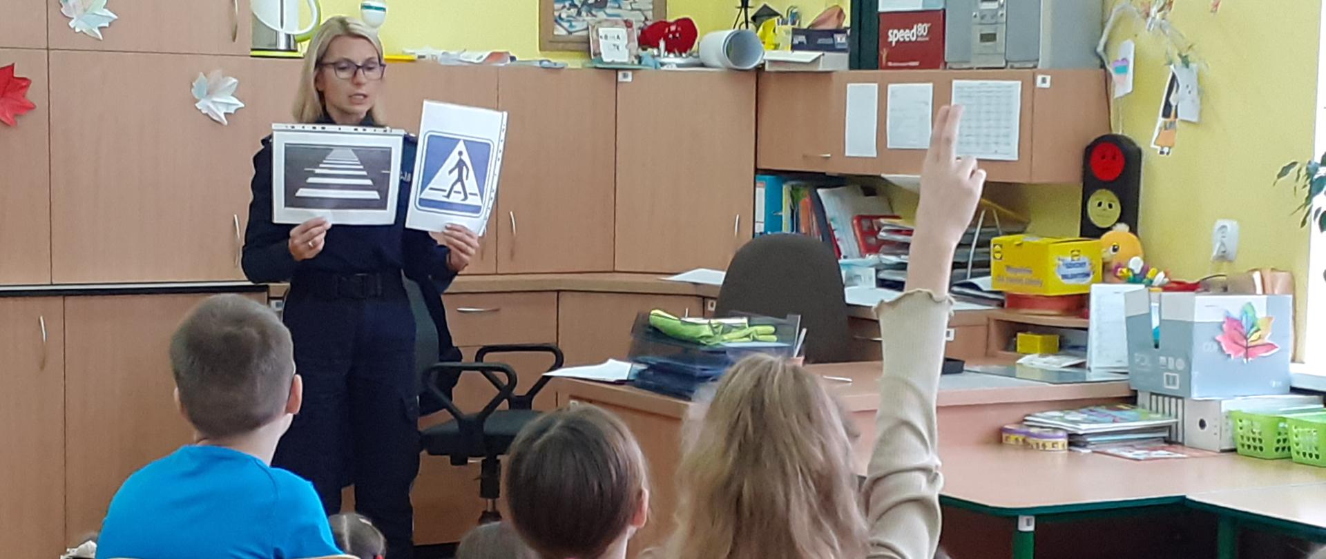 Zdjęcie policjantki trzymającej pomoce na tle szafek w świetlicy OMS I st. w Piszu. U dołu dzieci zgłaszające się do odpowiedzi.