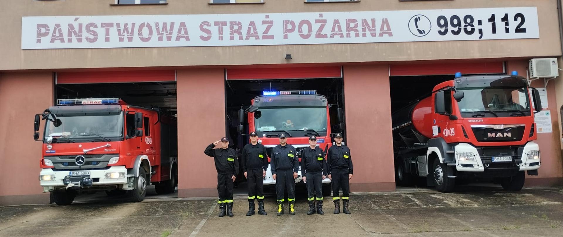 Przed remizą strażacką stoi kilku strażaków. Wrota garażowe są otwarte za nimi stoją czerwone wozy bojowe.