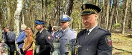 Uczestnicy uroczystości upamiętniających. Zdjęcie w ciągu dnia. Słonecznie. Widać las. Osoby umundurowane oraz w uraniach cywilnych.
