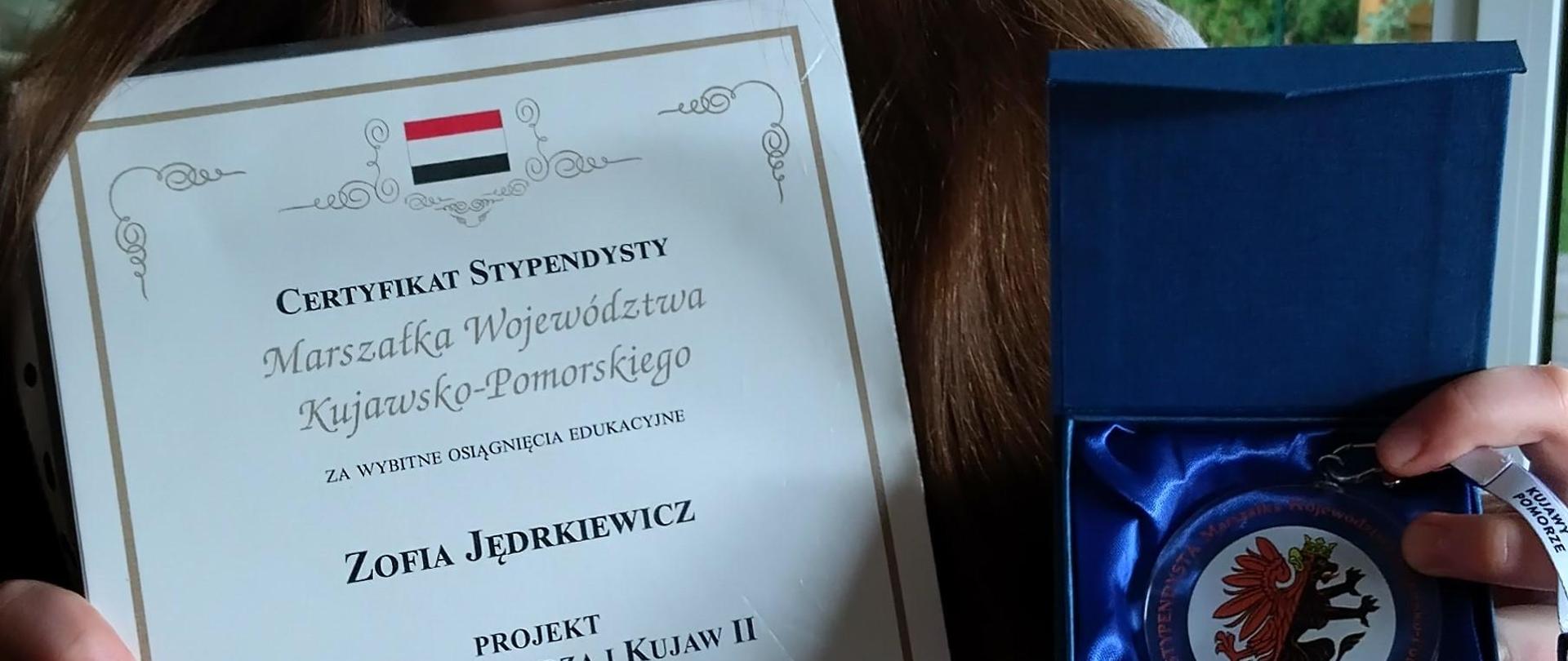 na zdjęciu w tle zielone krzewy za oknem na pierwszym planie ciemnowłosa dziewczyna trzymająca w rękach dyplom gratulacyjny i medal stypendysty 