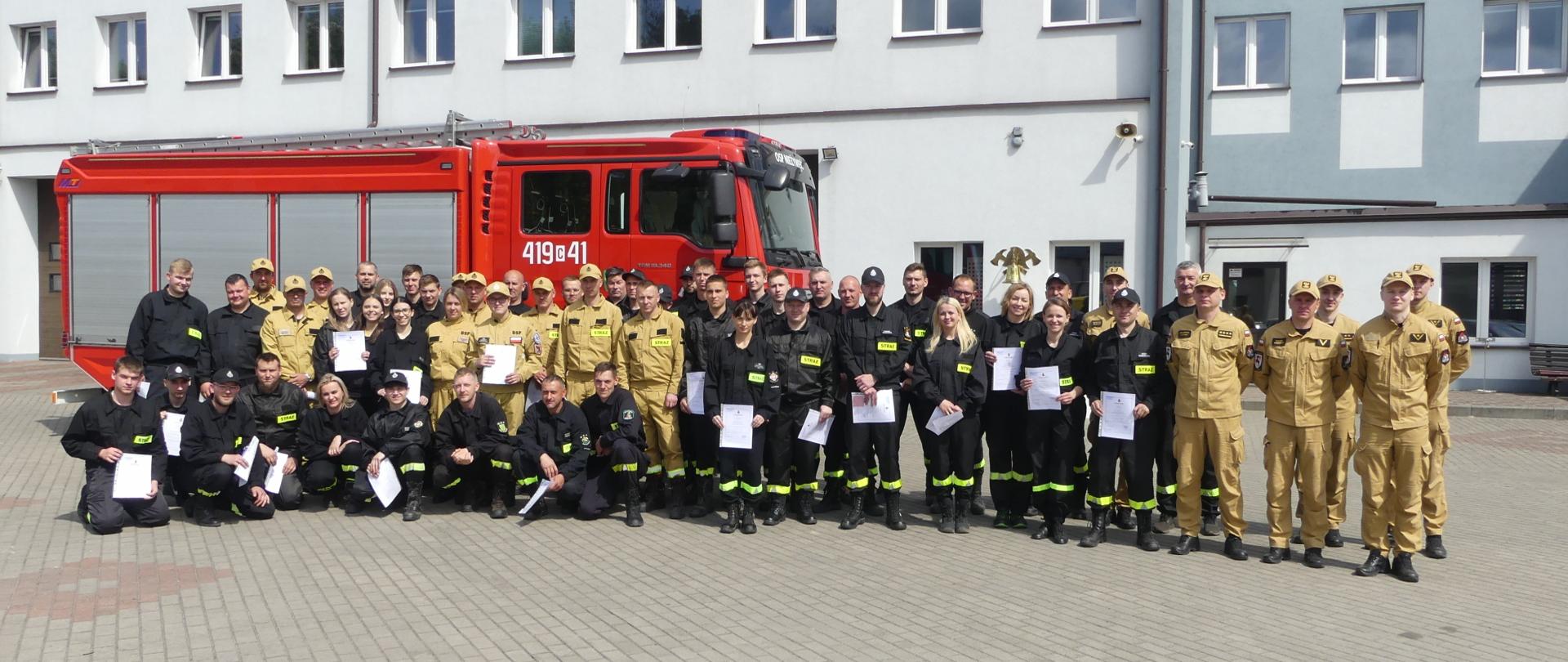 Zdjęcie przedstawia uczestników szkolenia podstawowego dla strażaków ratowników Ochotniczych Straży Pożarnych. Zdjęcie zbiorowe uczestników po ogłoszeniu pozytywnego wyniku egzaminu. Po prawo stoją funkcjonariusze Komendy Powiatowej. Za zgromadzonymi samochód gaśniczy a dalej budynek Jednostki Ratowniczo-Gaśniczej.