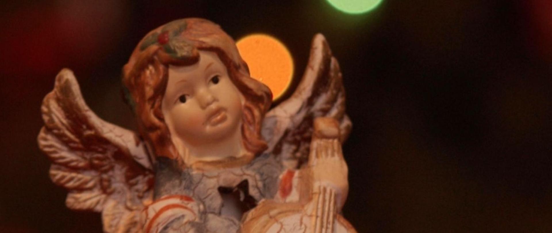 Plakat z figurką anioła trzymającego wiolonczelę stojącą na czerwonym obrusie. Na plakacie informacje o koncercie klasy wiolonczeli. 