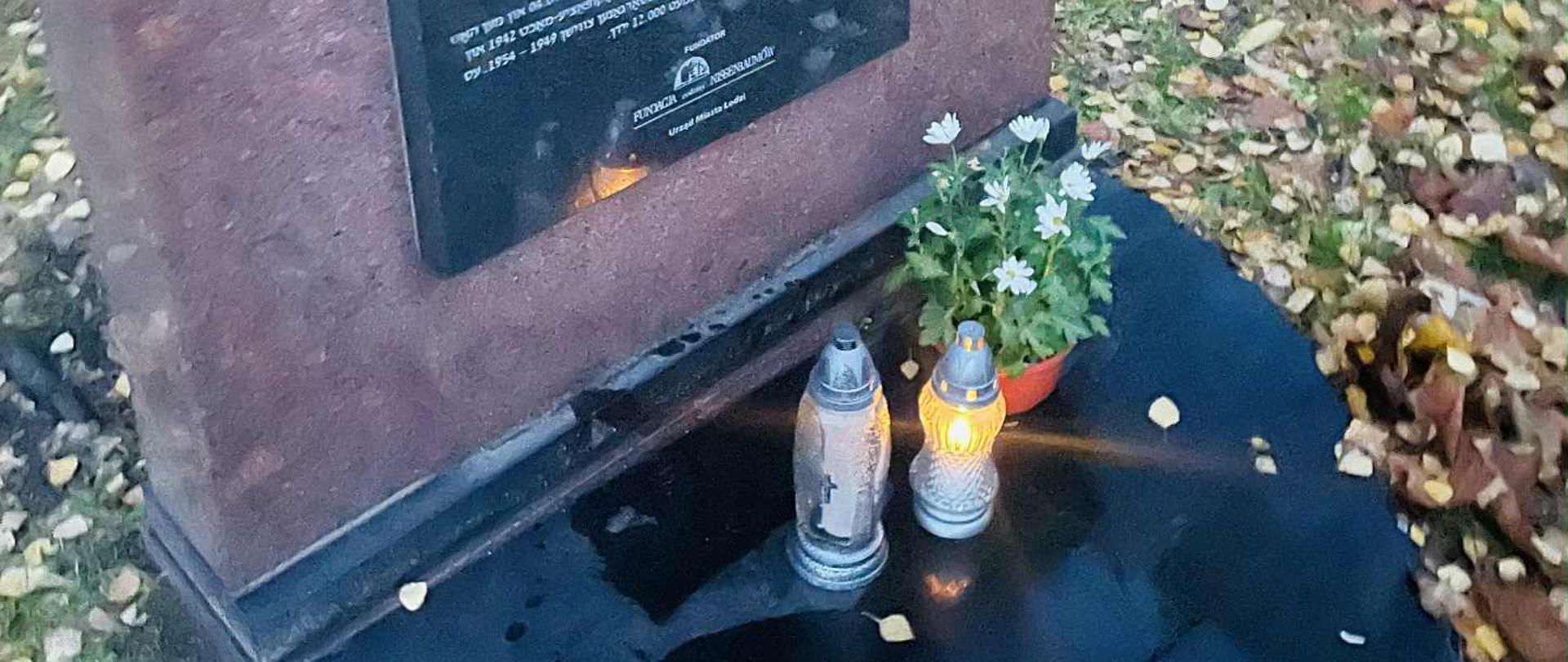 Na zdjęciu zapalone dwa znicze, białe kwiaty w doniczce oraz symboliczny kamień znajdujące się na macewie, żydowskim nagrobku, w formie pionowo ustawionej kamiennej, płyty.