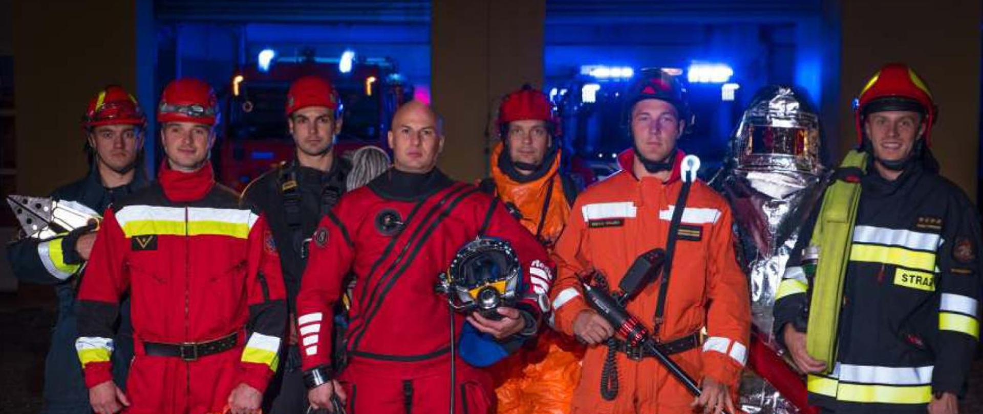 Zdjęcie przedstawia strażaków Państwowej Straży Pożarnej reprezentujących różne specyfiki działań prowadzonych przez formację. Strażacy ubrani są w różne umundurowanie oraz mają atrybuty reprezentujące ich specjalizację. Od lewej: strażak grupy specjalistycznej ratownictwa technicznego, strażak grupy specjalistycznej ratownictwa medycznego, strażak grupy specjalistycznej ratownictwa wysokościowego, strażak grupy specjalistycznej ratownictwa wodno-nurkowego, strażak grupy specjalistycznej ratownictwa chemiczno-ekologicznego, strażak grupy specjalistycznej poszukiwawczo-ratowniczej, strażak ubrany w ubranie żaroochronne oraz strażak w umundurowaniu specjalnym.