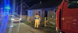 Na zdjęciu strażak w umundurowaniu specjalnym z kamizelką z napisem „Kierujący Działaniem Ratowniczym”. Strażak przed budynkiem mieszkalnym, w którym wcześniej panował pożar. Na zdjęciu dodatkowo widoczne samochody ratowniczo-gaśnicze oraz radiowóz policyjny.