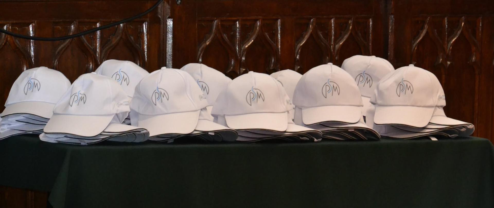 Rząd białych czapeczek z logo PSM ułożonych na zielonym suknie stołu w Sali Królewskiej, przygotowanych dla wyróżnionych uczniów podczas zakończenia roku szkolnego.