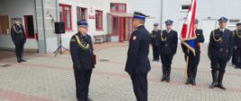 Zastępca pomorskiego komendanta wojewódzkiego Państwowej Straży Pożarnej stoi z czerwoną teczką przed nim stoi strażak obok poczet sztandarowy Komendy Powiatowej Państwowej Straży Pożarnej w Kwidzynie. 