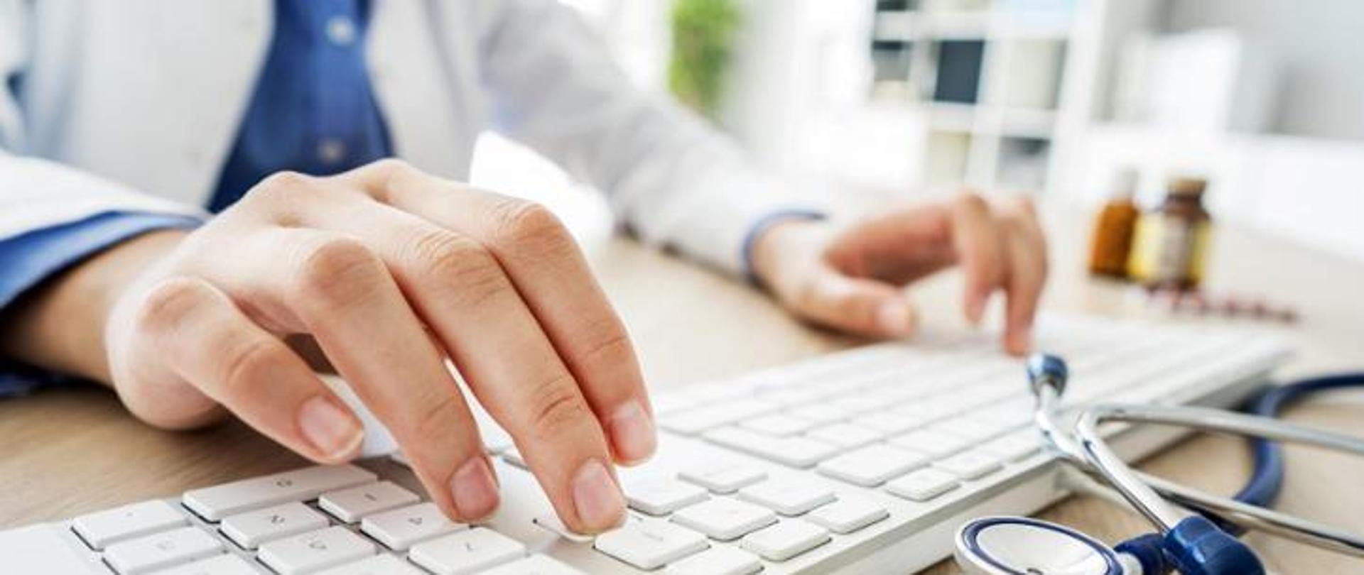 zdjęcie dłonie lekarza na klawiaturze, na pierwszym planie stetoskop