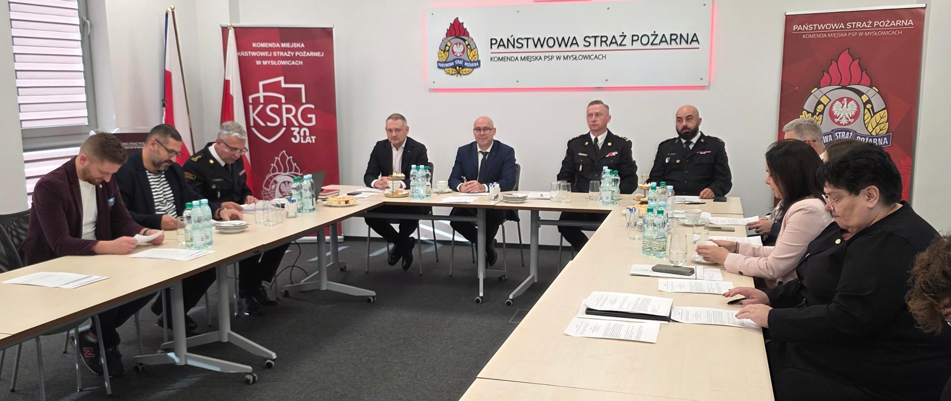 Zdjęcie przedstawia spotkanie komitetu założycielskiego, która odbywa się w sali konferencyjnej Komendy Miejskiej Państwowej Straży Pożarnej w Mysłowicach.
Uczestnicy: Wokół długiego, prostokątnego stołu konferencyjnego (w kształcie litery "U" lub dwóch połączonych stołów) siedzi kilkanaście osób, głównie w strojach cywilnych, ale również w mundurach.
W środkowej części, po prawej stronie stołu, siedzą trzej mężczyźni w eleganckich mundurach służbowych (jeden w ciemnym, a dwaj w jasnym kolorze khaki z naszywkami i pagonami), co sugeruje ich rangę i funkcję w Straży Pożarnej.
Pozostali uczestnicy są ubrani w odzież cywilną, m.in. garnitury, koszule i marynarki, a także kilka kobiet siedzi po prawej stronie stołu.
Wszyscy uczestnicy mają przed sobą dokumenty, notatniki oraz butelki z wodą.
Wystrój sali: Tło jest jasne, utrzymane w kolorystyce bieli i szarości.
Na ścianie za stołem wiszą dwa duże, czerwono-białe banery/plakaty. Centralny plakat zawiera logo i napis: "PAŃSTWOWA STRAŻ POŻARNA KOMENDA MIEJSKA PSP W MYSŁOWICACH".
Po lewej stronie znajduje się drugi plakat z logo KSRG (Krajowego Systemu Ratowniczo-Gaśniczego) i napisem "KSRG 30 LAT".
W lewym rogu widoczny jest stojak z flagą Polski.
Zdjęcie dokumentuje oficjalne spotkanie Komitetu Założycielskiego Sztandaru dla KM PSP w Mysłowicach z udziałem kadry kierowniczej PSP w Mysłowicach oraz, przedstawicieli władz lokalnych lub innych służb.