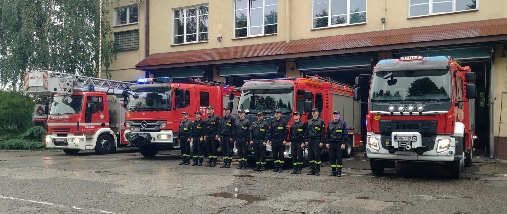 Cztery samochody pożarnicze stoją przed garażami JRG. Przed pojazdami stoi w szeregu zmiana służbowa JRG Wadowice.