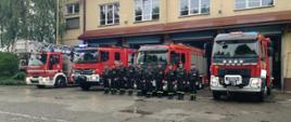 Cztery samochody pożarnicze stoją przed garażami JRG. Przed pojazdami stoi w szeregu zmiana służbowa JRG Wadowice.
