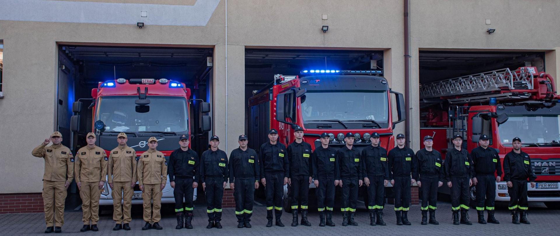 Zdjęcie przedstawia strażaków stojących przed budynkiem strażnicy przed samochodami pożarniczymi oddając hołd strażakom.