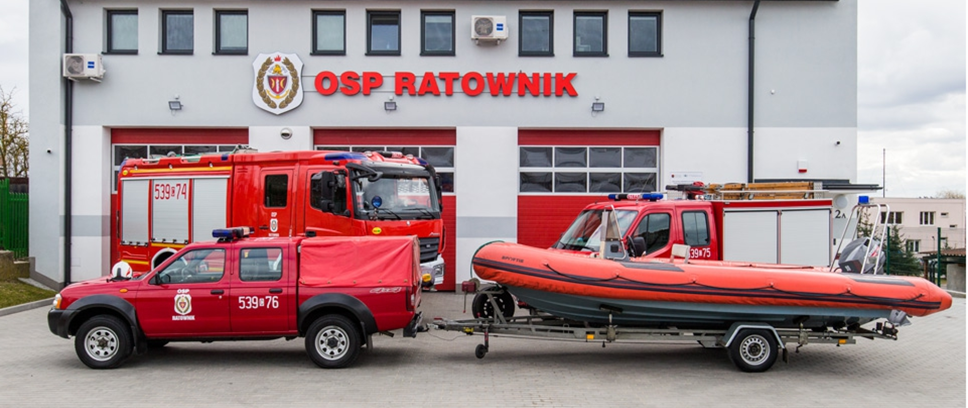 Przed bramami jednostki Ochotniczej Straży Pożarnej Ratownik stoi bokiem skierowany w lewą stronę samochód pożarniczy Nissan z podczepiona przyczepką na której znajduje się łódź hybrydowa, za nimi skierowane do siebie po lewej stoi samochód gaśniczy Mercedes a po prawej Renault.