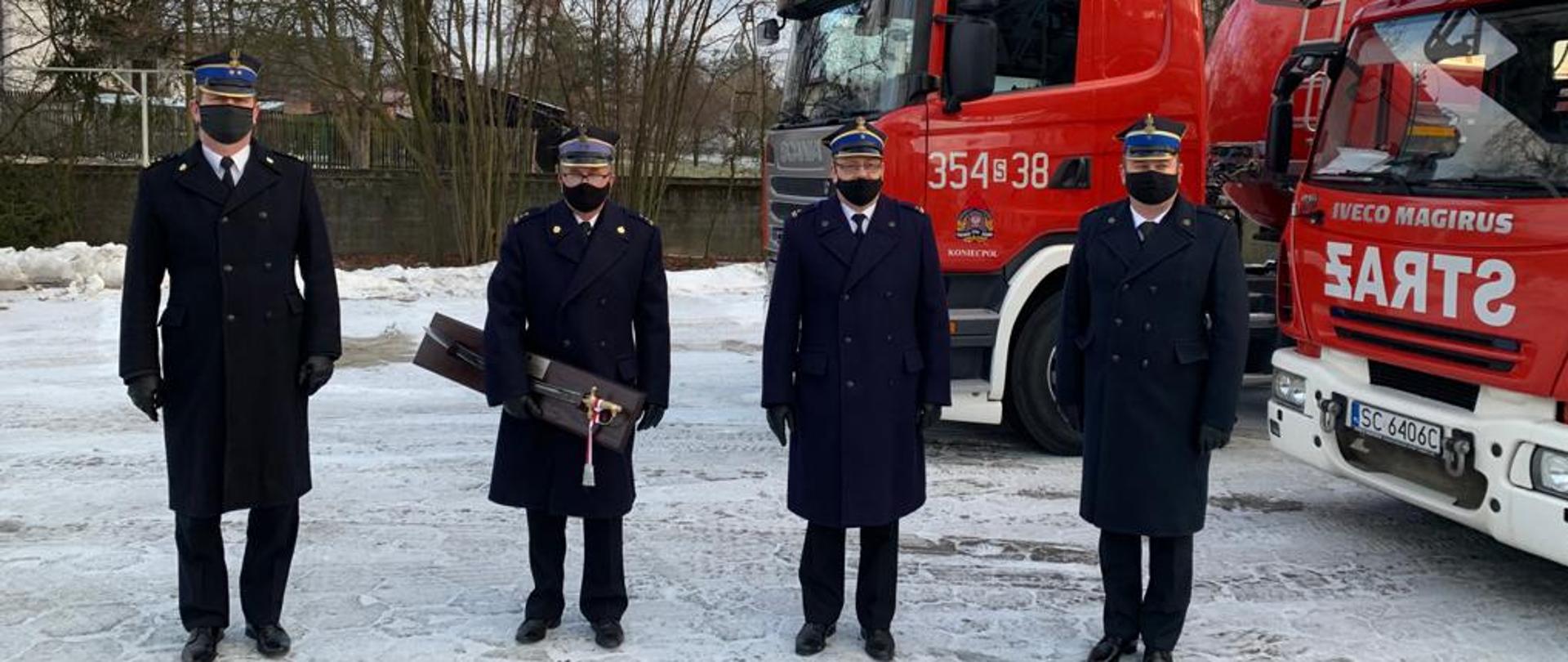 Zdjęcia przedstawia 4 funkcjonariuszy Państwowej Straży Pożarnej w mundurach wyjściowych na tle samochodów pożarniczych. 