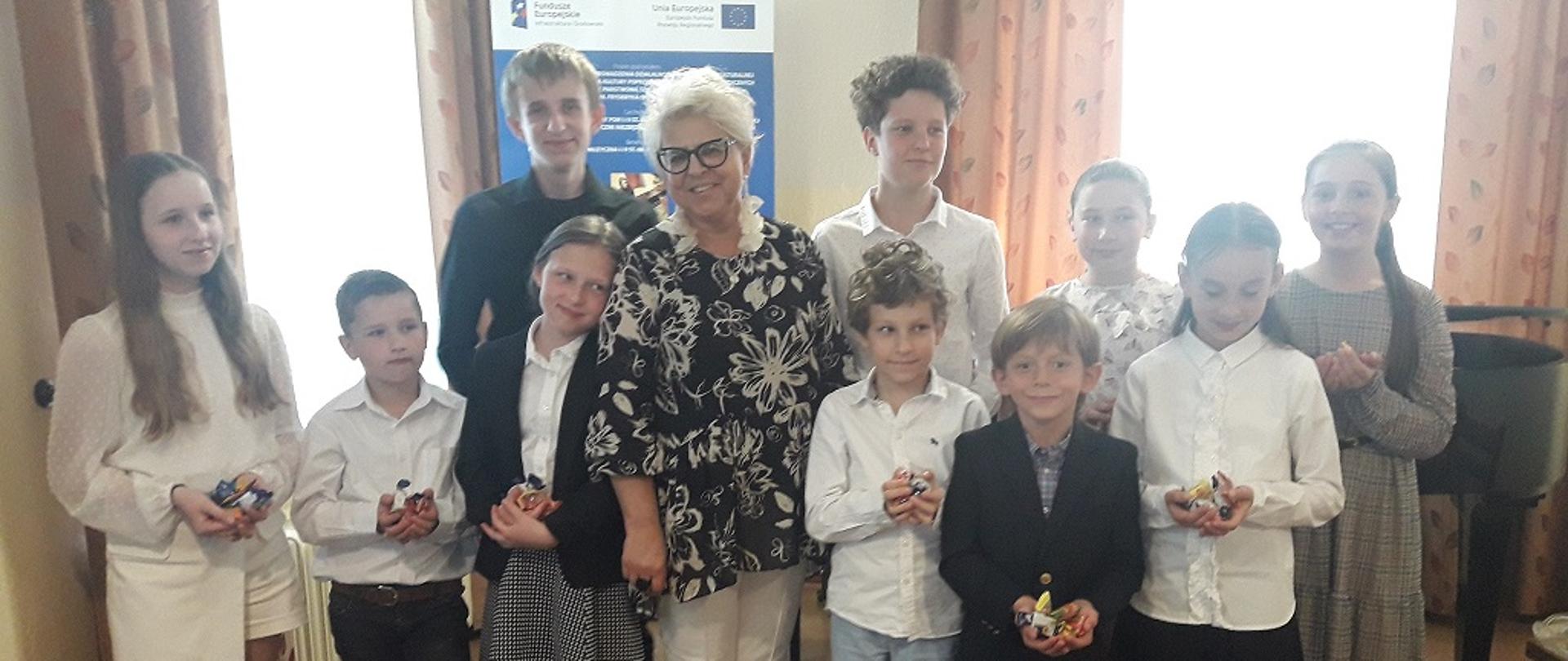 Zdjęcie uczniów klasy fortepianu po audycji pianistycznej. Stoją wraz z nauczycielką w dwóch rzędach, za nimi baner unijny, dwa okno przysłonięte firaną i zasłonami, z prawej strony fortepian, na pierwszym planie puste krzesła
