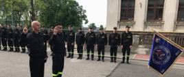 Fotografia została wykonana na placu komendy, po prawej stronie widać sztandar komendy, naprzeciwko sztandaru stoi dwóch strażaków w trakcie ślubowania. W tle widnieją pozostali strażacy ustawieni w rzędzie.
