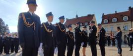 Strażacy stoją w rzędzie na środku placu apelowego. Komendanci wręczają im awanse na wyższe stopnie służbowe. W tle dwie kamienice i inni strażacy. 