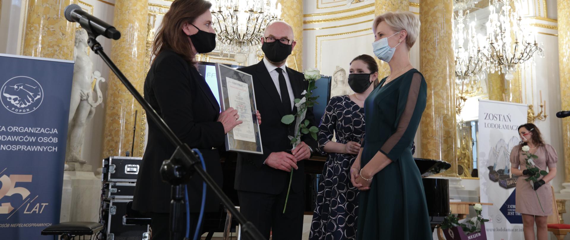 Na zdjęciu trzy kobiety i mężczyzna. Jedna z kobiet wręcza drugiej dyplom w ramce. Mężczyzna trzyma w ręku białą różę.