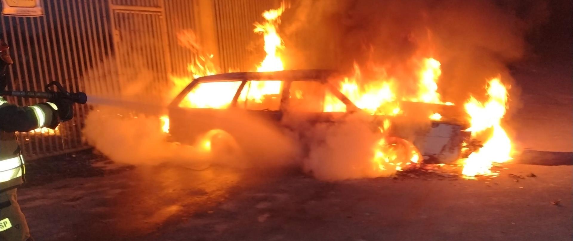 Płonące w całości auto na poboczu drogi przy ogrodzeniu. Strażak z prądownicą prowadzi działania gaśnicze. Pora nocna.