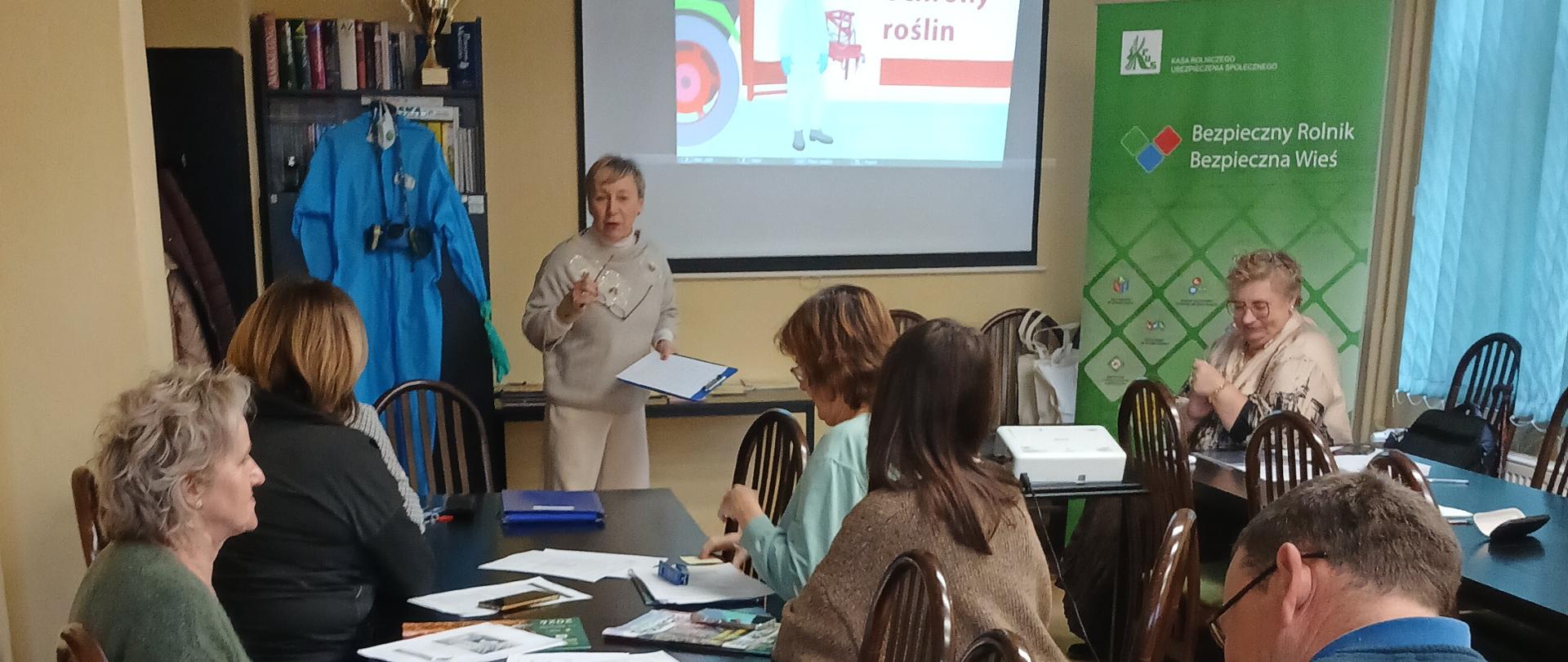 Uczestnicy spotkania przy stole konferencyjnych, w tle ekran z prezentacją i prelegent w trakcie wykładu