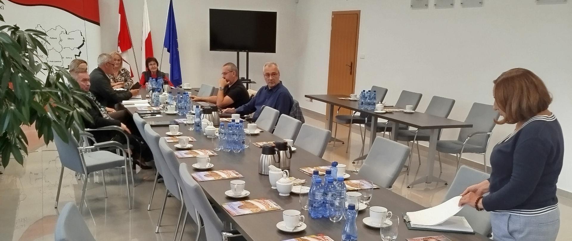 Sala konferencyjna z długim stołem nakrytym filiżankami, butelkami wody i materiałami informacyjnymi, przy którym siedzi kilka osób podczas spotkania; w tle widoczne są flagi Polski i Unii Europejskiej oraz godło państwowe.