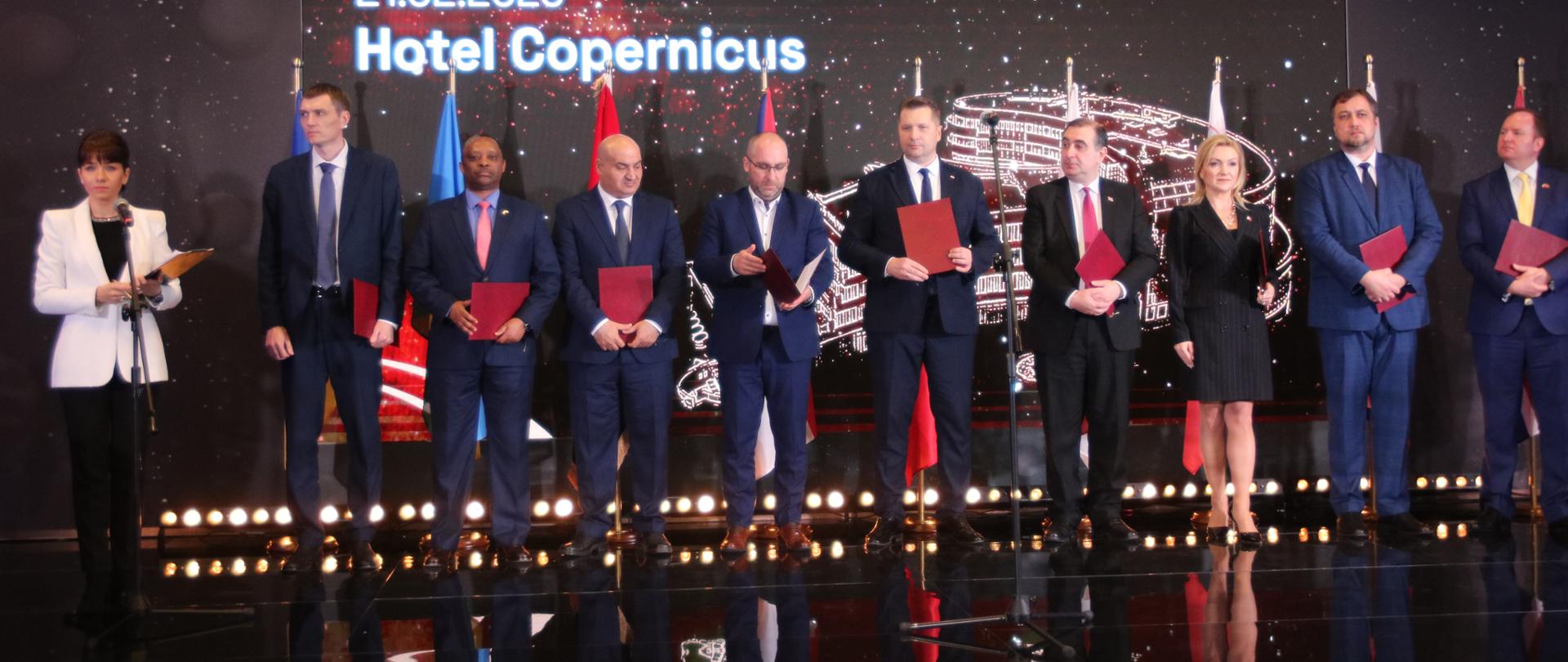 Kilka trzymających czerwone teczki osób stoi na błyszczącej podłodze, za nimi na ścianie napis 21.02.2023 Hotel Copernicus, z boku stoi ubrana na biało kobieta i mówi do mikrofonu na stojaku.