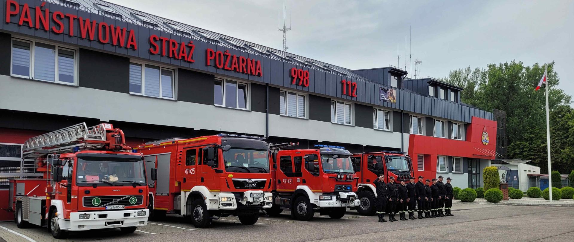 Zdjęcie zrobione w dzień. Na zdjęciu widać strażaków stojących przed samochodami strażackimi i w oddali widać budynek jednostki. 