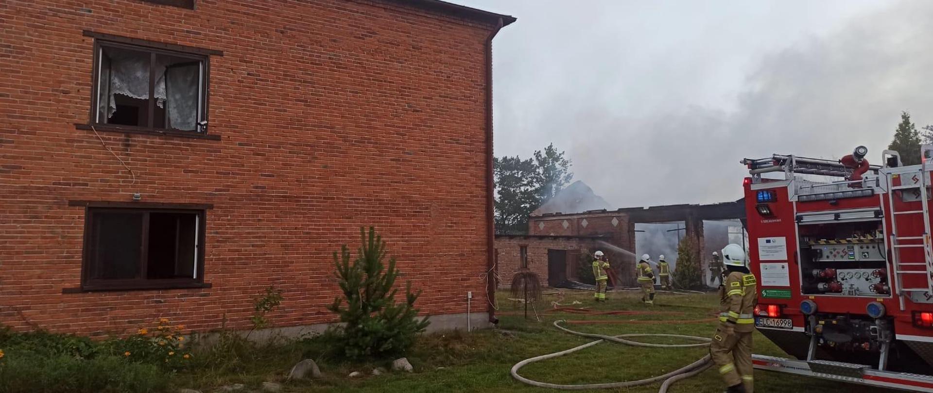 Na zdjęciu widać strażaków w żółtych strojach i białych hełmach, którzy gaszą pożar budynku murowanego z czerwonej cegły. Budynek nie ma dachu oraz drzwi. Unosi się nad nim dym. Na trawiastym podwórku stoi czerwony samochód strażacki oraz ułożone są węże strażackie czerwone i białe. Obok jest dom, w którym otwarte są okna. W tle widać drzewa. Jest widno.