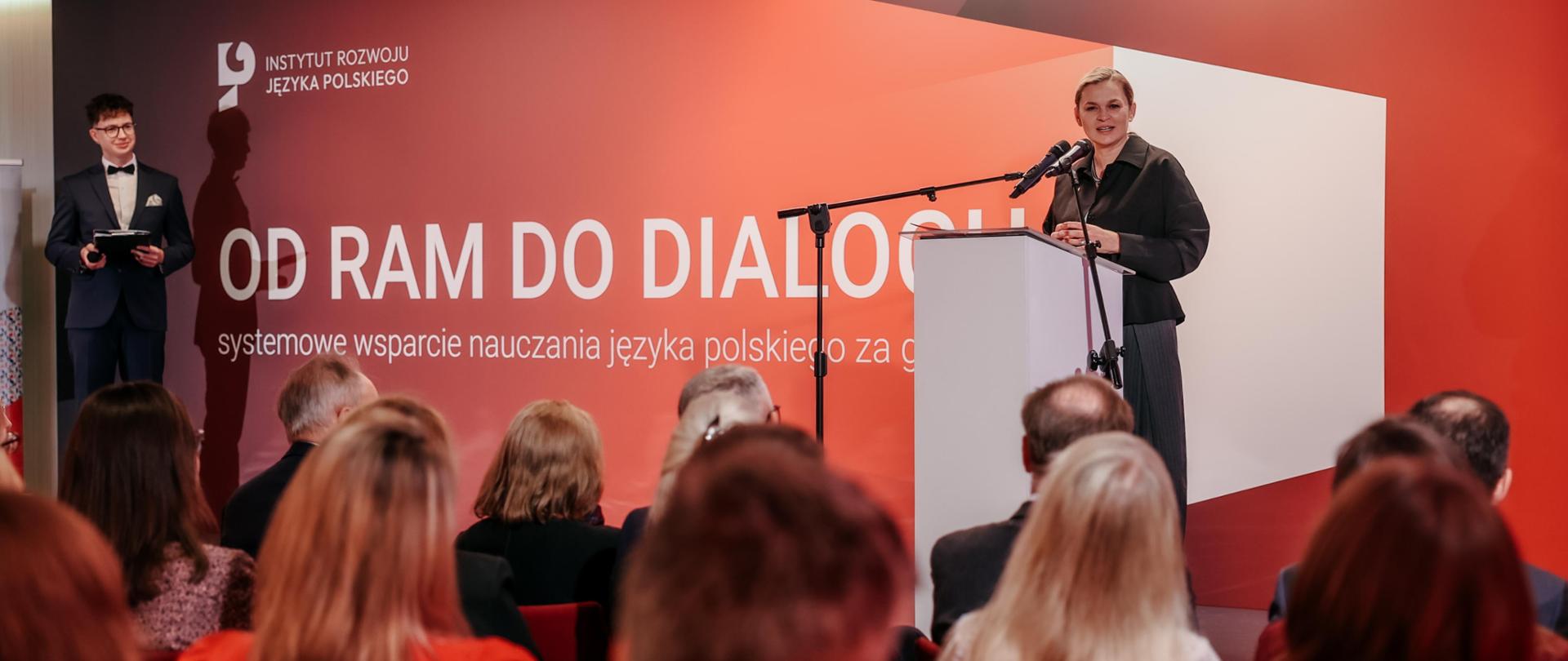 Ujęcie z sali konferencyjnej. Minister Edukacji Barbara Nowacka stoi za mównicą na scenie i przemawia do publiczności. Za nią widać duży, czerwono-biały baner z hasłem "OD RAM DO DIALOGU. systemowe wsparcie nauczania języka polskiego za granicą".